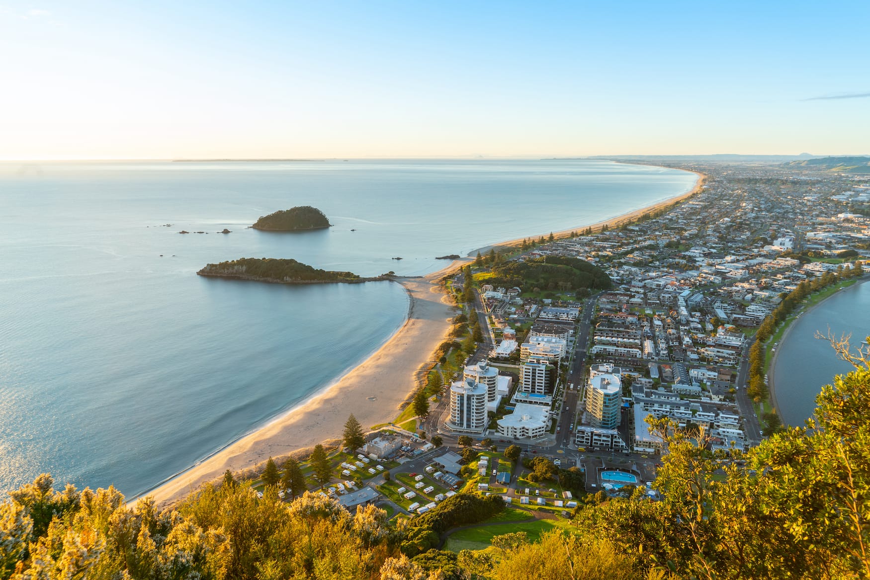 Der Mount Maunganui erstreckt sich unter uns, während die Sonne am Horizont aufgeht und über den Strand und die Gebäude am Meer fällt
