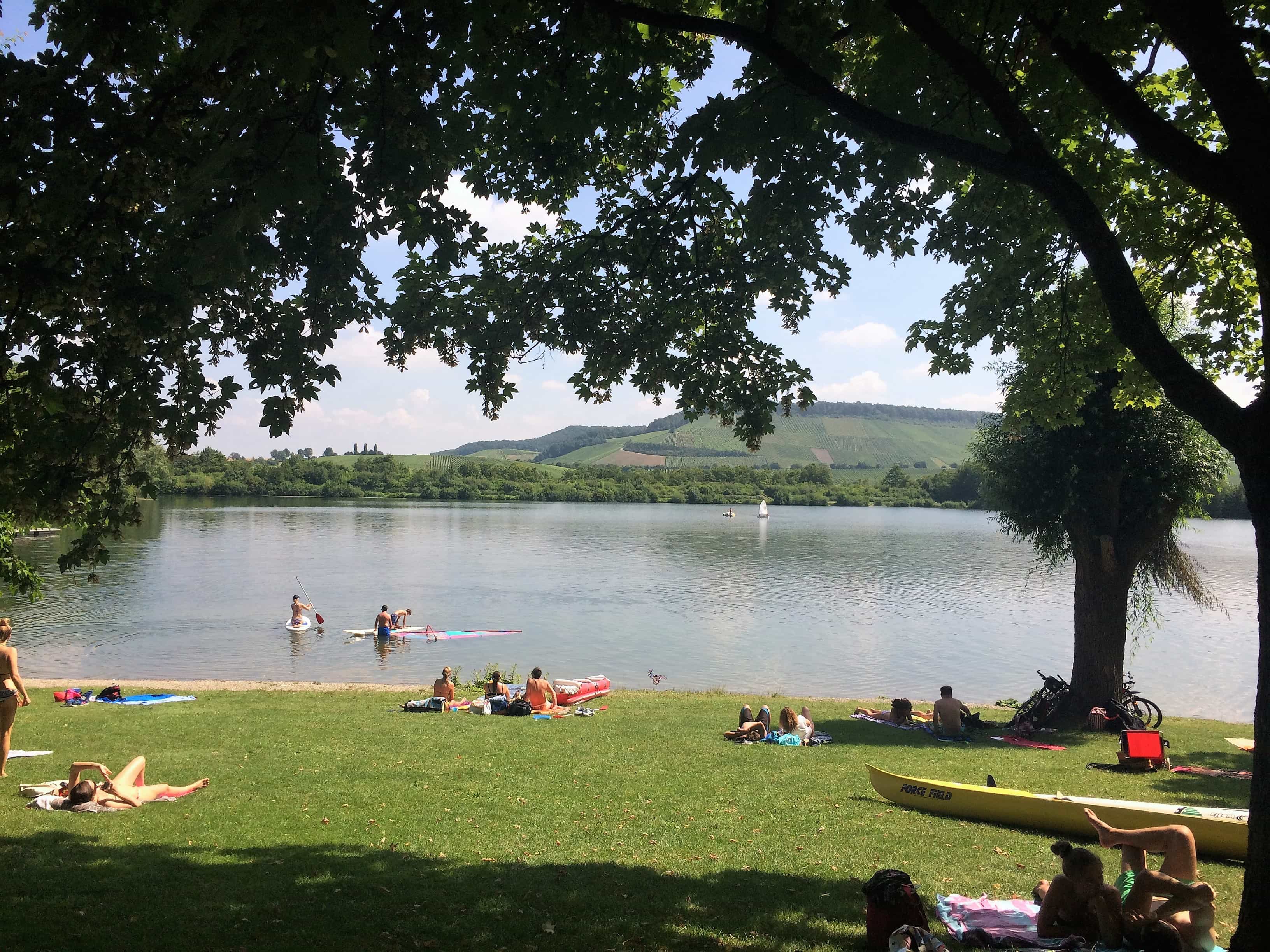 Sommertag am Breitenauer See; einige Menschen liegen auf Handtüchern am Ufer, im Wasser ist eine Gruppe Menschen mit Stand-up-Paddle-Boards unterwegs.