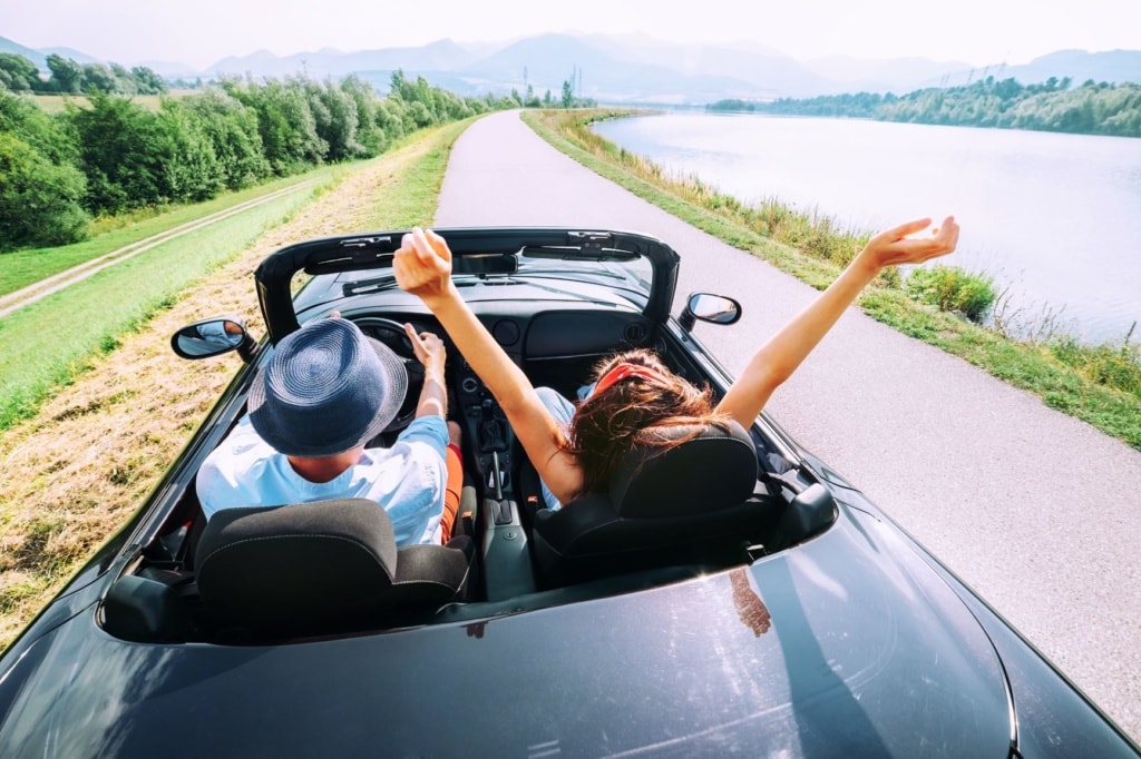 Mann und Frau fahren im Cabrio parallel zu einem Fluss 
