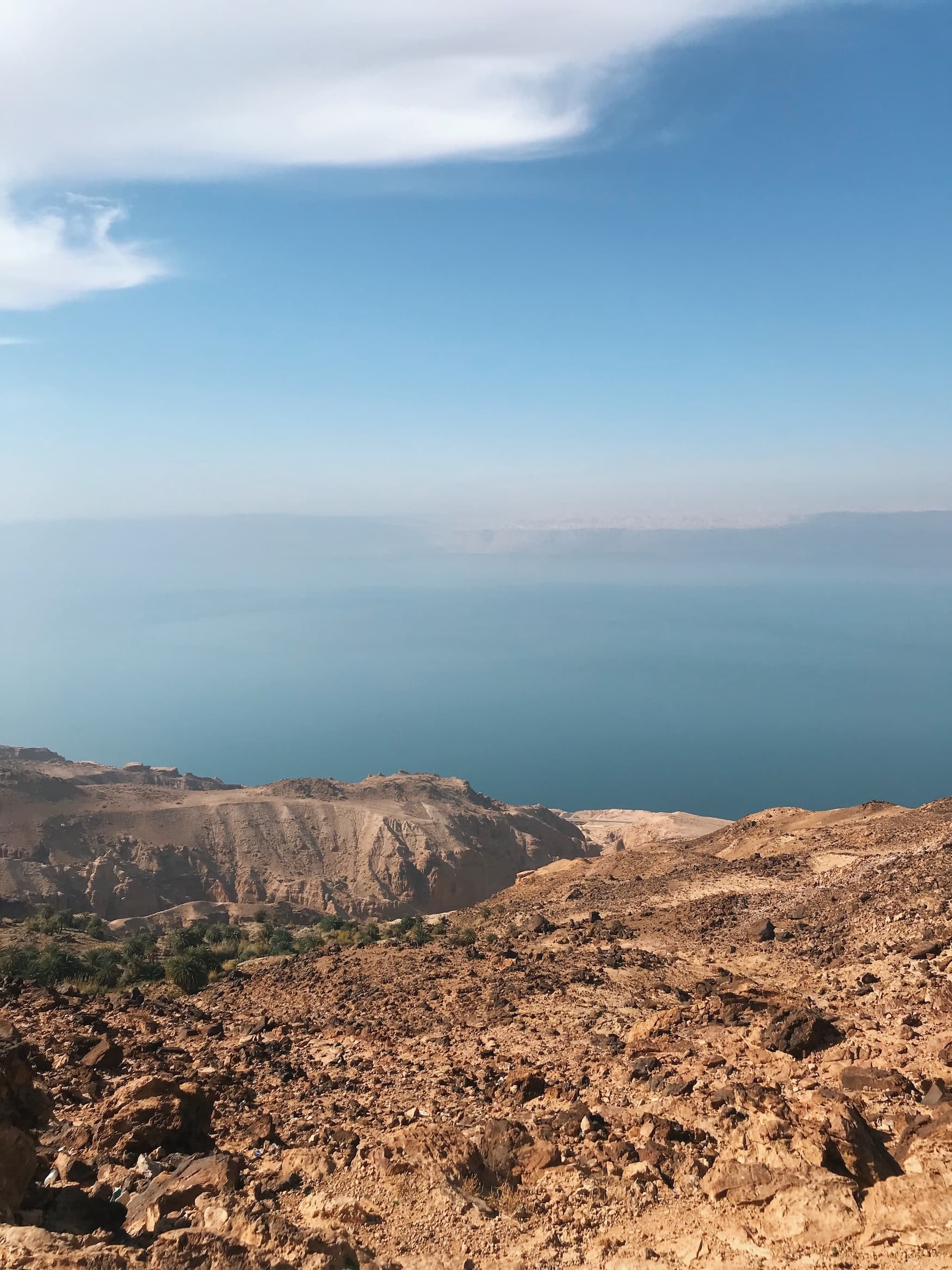 Im Vordergrund ine trockene, schroffe Landschaft und im Hintergrund kann man das türkisfarbene Wasser des Toten Meers erahnen