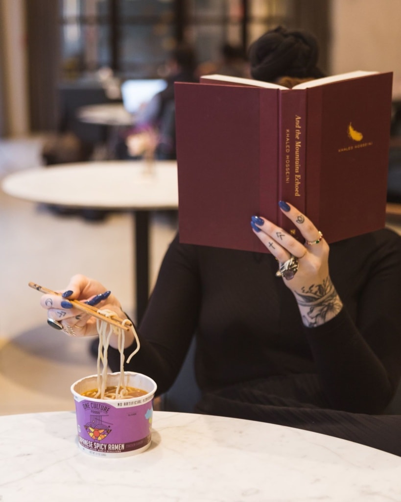 Frau hinter Buch sitzt am Tisch, liest und isst Instantnudelsuppe