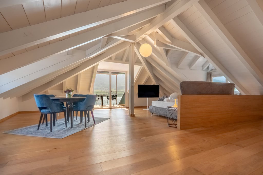 Hotelzimmer im Dachgeschoss, der Boden mit Dielen verkleidet, links eine kleine Sitzgruppe, rechts ein Doppelbett, auf einen Fernseher ausgerichtet