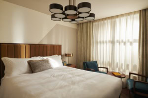 Ein Hotelzimmer im Mid-Century-Stil, mit breitem Bett, mit Samt gepolstertem Kopfteil und einer schwarzen Kunststoff-Designlampe an der Decke.