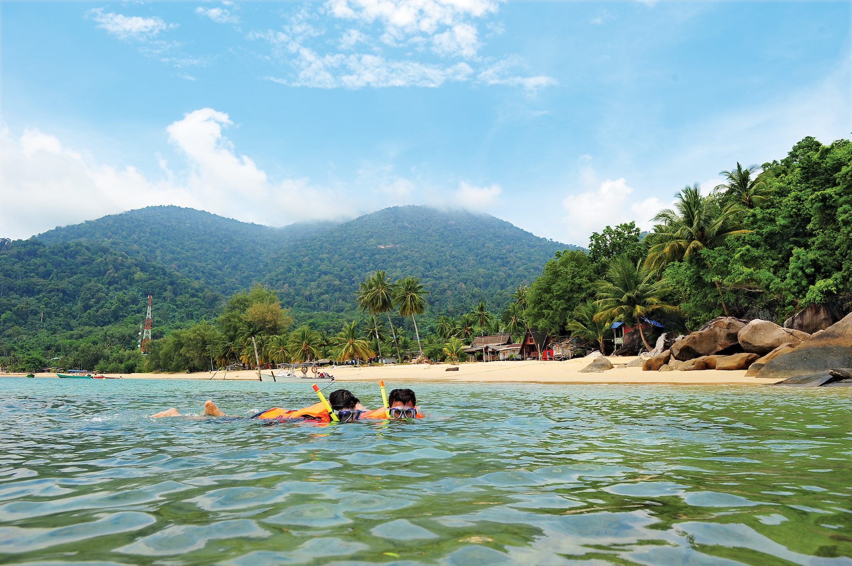 Im strandnahen Meer einer von dichtem Wald bewachsenen Tropeninsel mit mehreren Bergen schwimmen zwei Menschen mit Schnorchelbrillen flach im Wasser. Am Sandstrand hinter ihnen liegen zur Rechten einige Felsen, mittig hinter den beiden Tauchern ragen einige kleine Hütten aus dem ufernahen Wald heraus. Links am Ufer steht ein kleiner, rot und weiß gestrichener Funkturm.