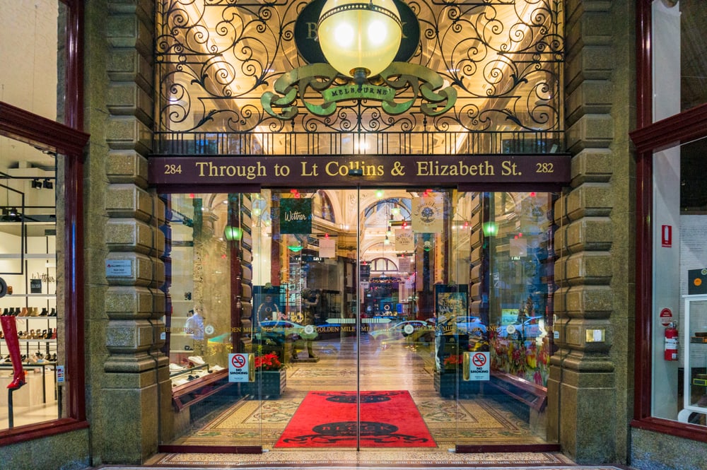 Eingang zu einer noblen Einkaufshalle: Im Türrahmen hängt über der doppelflügigen Glastür ein Querbalken mit der Aufschrift "Thoru to Lt Collings & Elizabeth Str. 282". Die Passage dahinter besteht aus eleganten Bodenfliesen, hohen Wänden und einer hell erleuchteten Decke. Links und rechts davon Geschäfte.