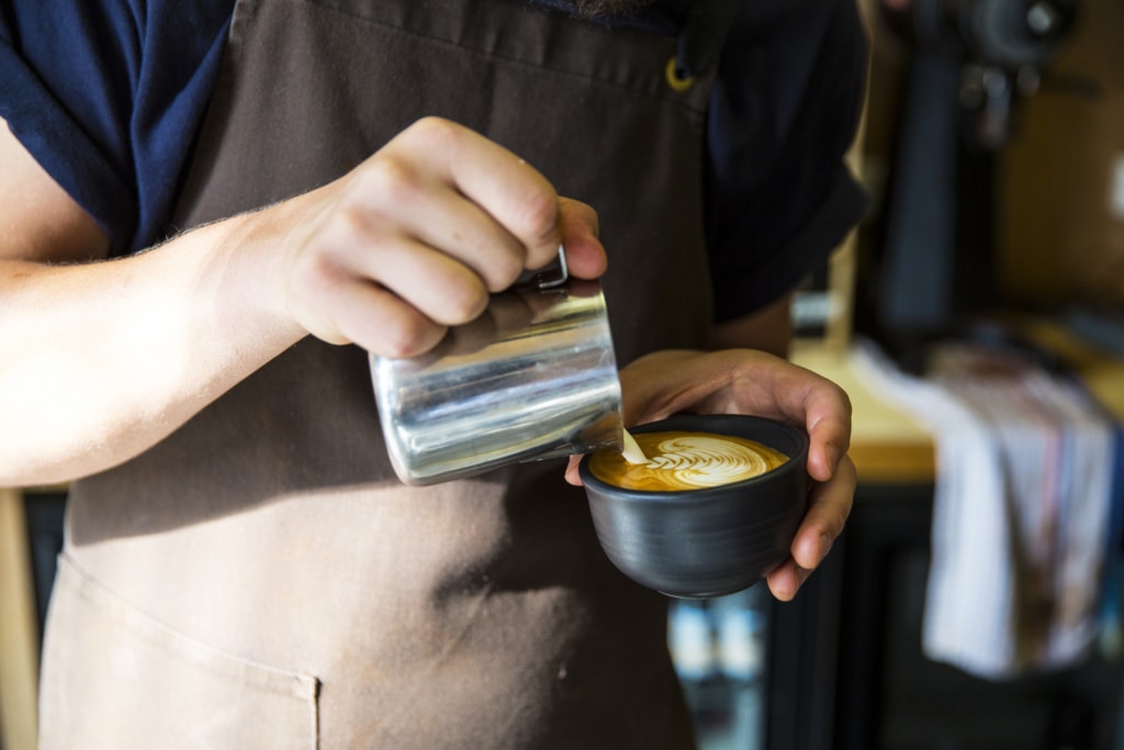 Ein Barrista mit brauner Schürze schüttet Milchschaum in eine schwarze Tasse mit Espresso und formt ein Muster. Sein Gesicht ist nicht zu erkennen.