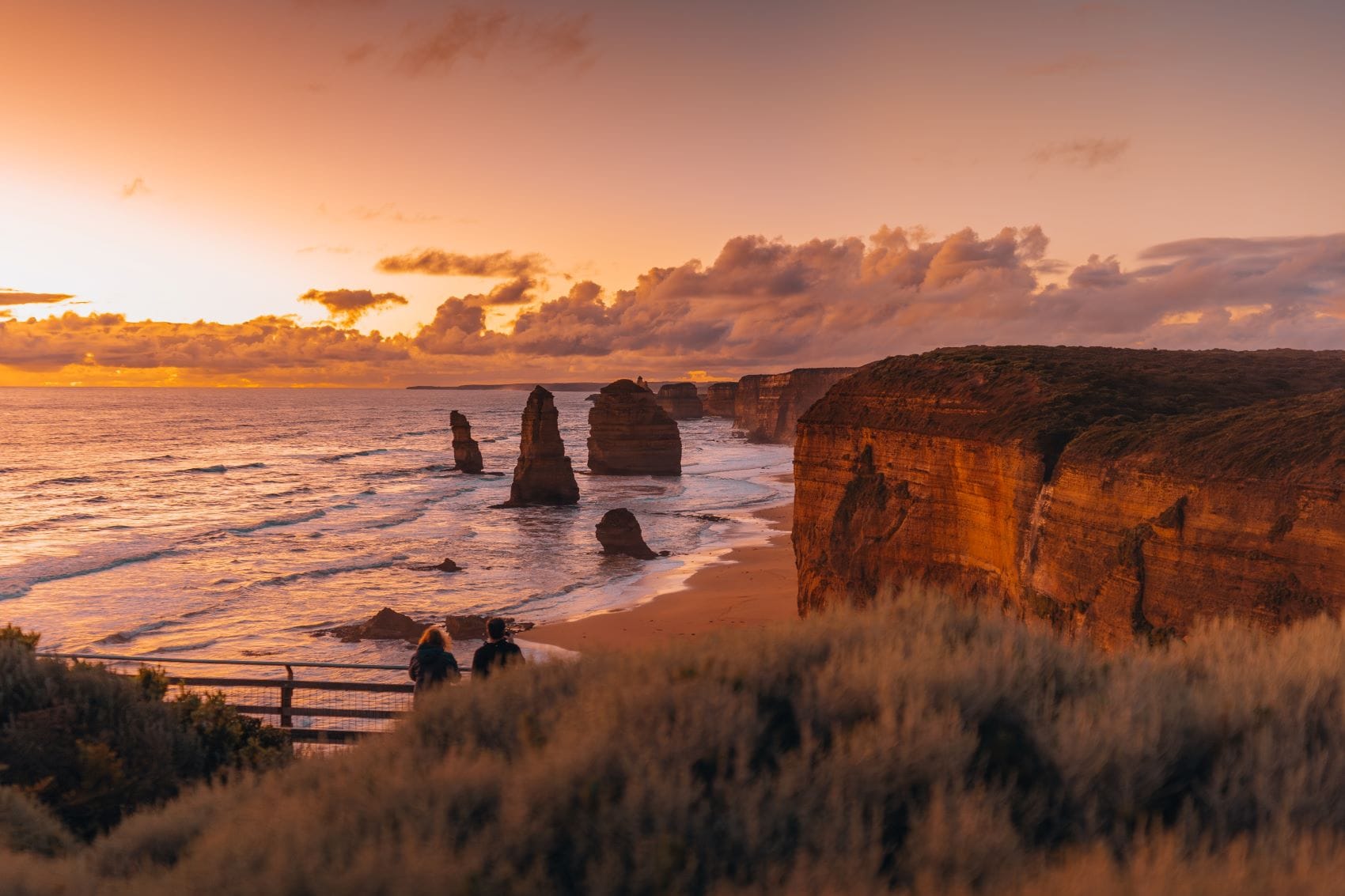 Vor einer Steilklippe auf der rechten ragen drei sehr hohe und mehrere niedrige, aber alle sehr schmale Felsen aus dem Meer. Die ganze Szene wird von der Sonne rot angestrahlt. Hinter einer Düne im Vordergrund lugen zwei Menschen hervor, die die Felsen betrachten.