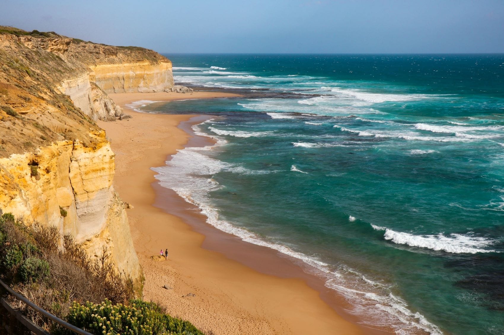 Blick von einer sandigen Klippe auf einen Strand: rechts das Meer, das an den mittigen Strand braust, links die Fortführung der Sandklippen, auf denen der Betrachter steht. Sie ziehen sich bis zum himmelblauen Horizont.