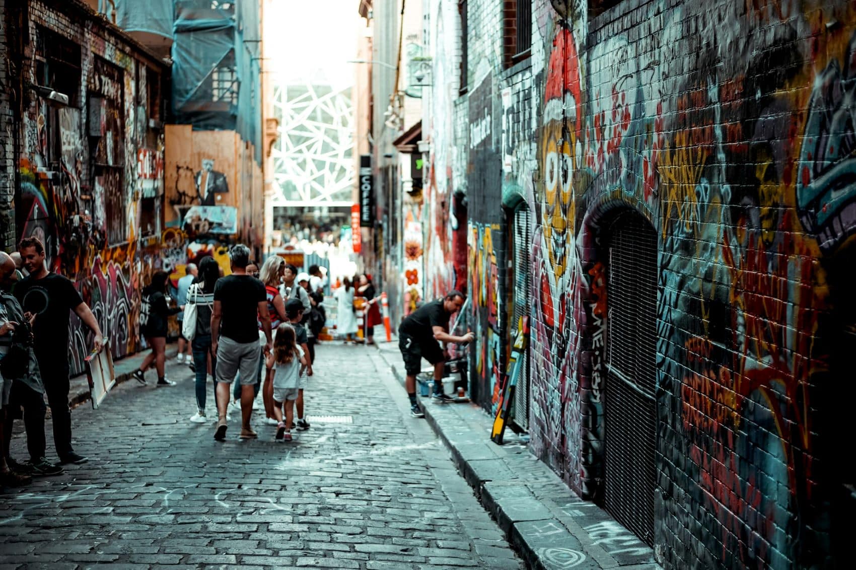 Eine gepflasterte Gasse mit Ziegelgebäuden zu beiden Seiten. Die Wände sind über und über mit Graffiti besprüht, viele Menschen sind in der Gasse unterwegs.