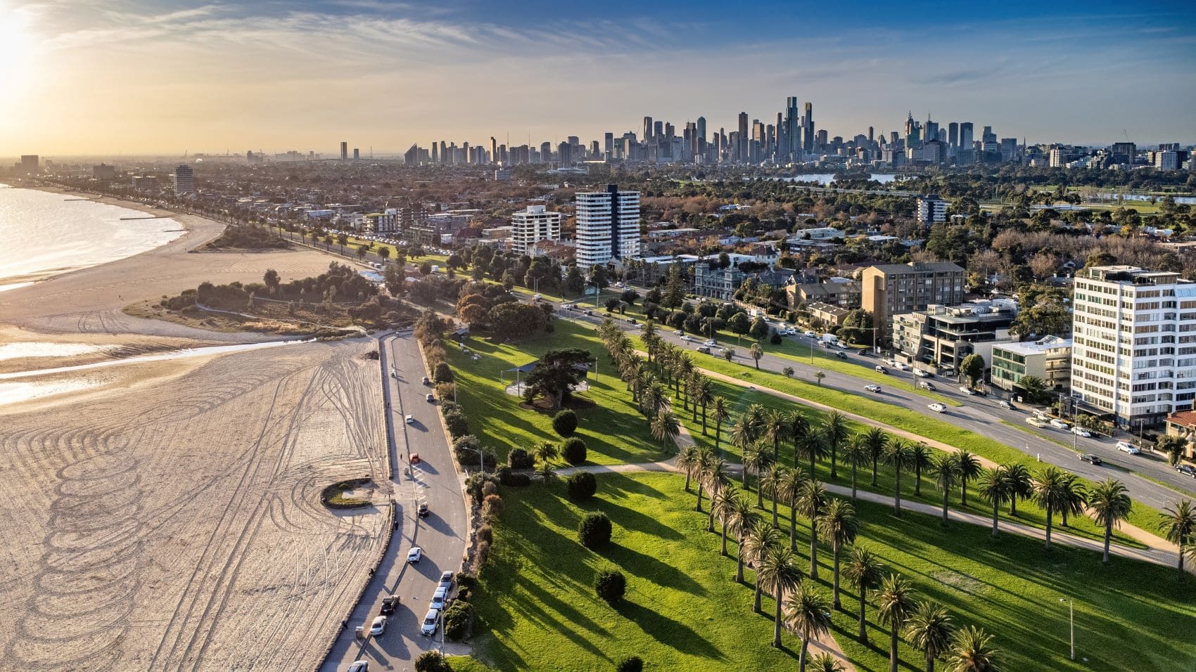 Drohnenaufnahme eines Stadtstrandes; links das Meer, dann ein grüner Parkstreifen, dann rechts die Ausläufer der Stadt, einige moderne Wohnhäuser. Weit im Hintergrund ist eine breite Skyline zu erkennen.