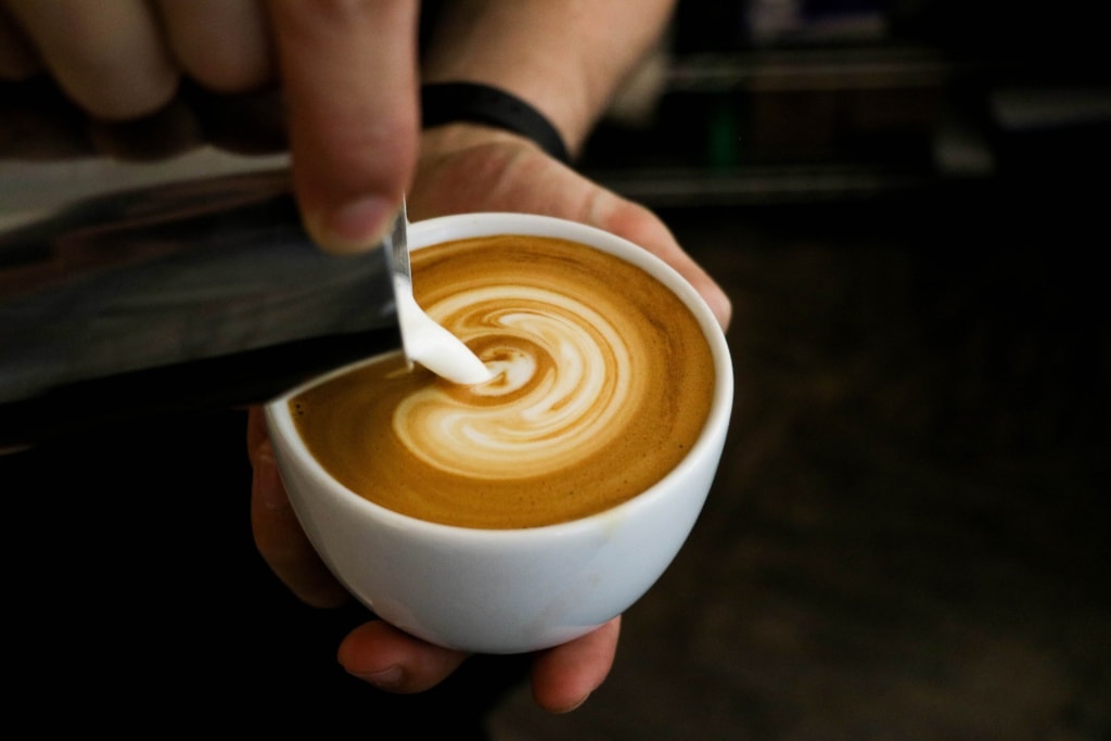 Person hält Kaffeetasse in der Hand und schüttet mit der anderen geschäumte Milch hinein