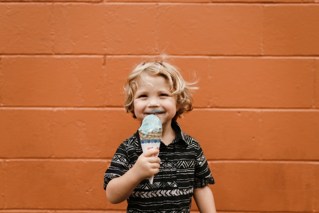 Ein kleiner Junge steht vor einer orangefarbenen Wand und schleckt ein Eis.