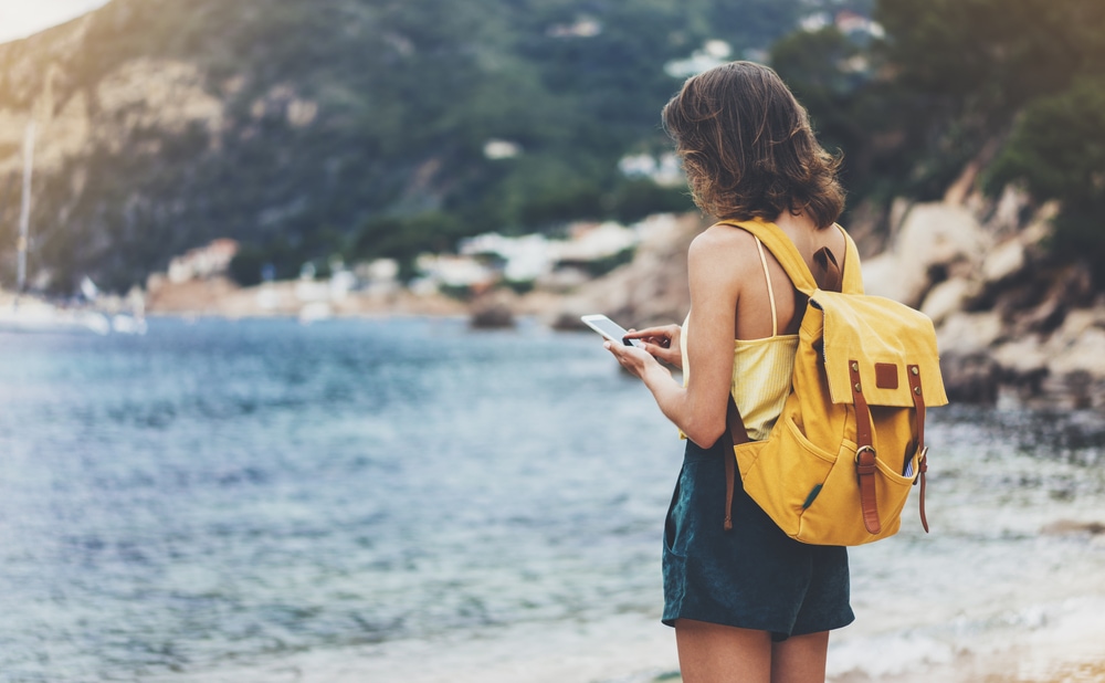Junge Frau steht am Meer und tippt ins Handy