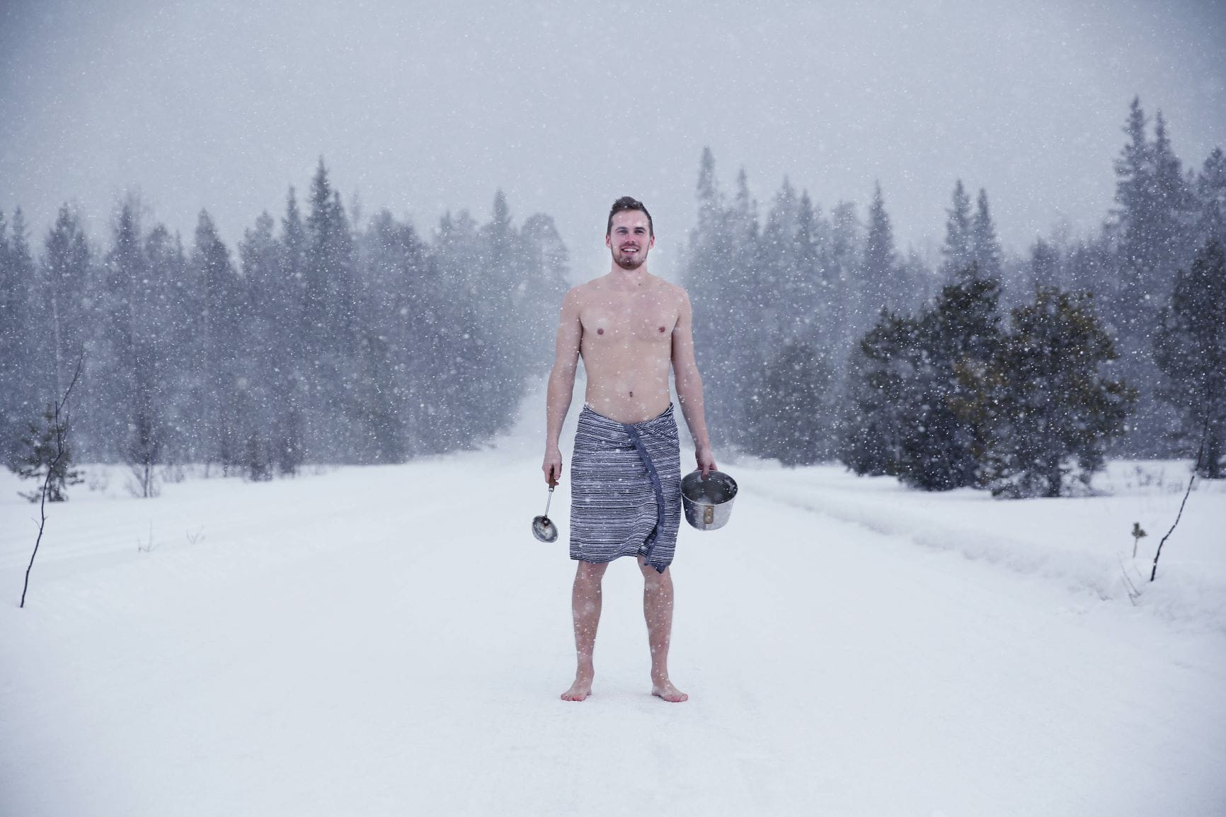 Mann steht halbnackt mit Sauna-Utensilien in finnischer Winterlandschaft