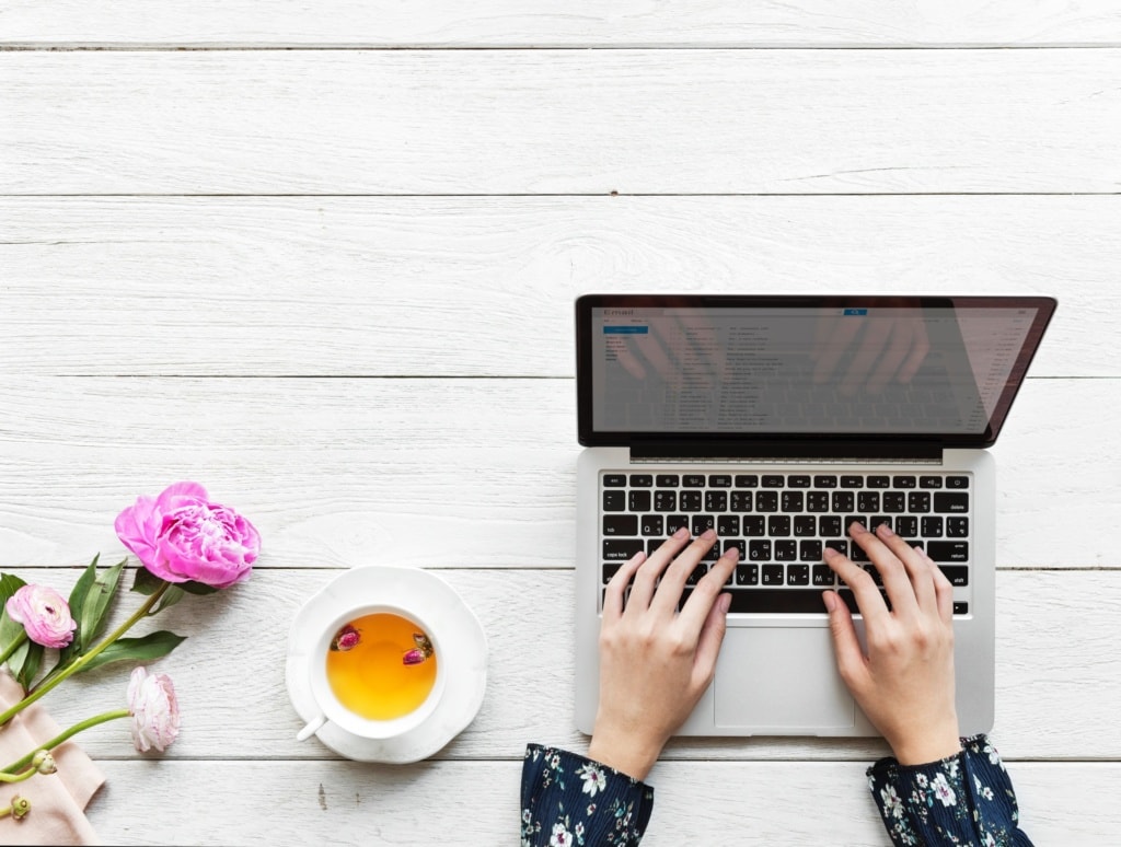 Person tippt auf Tastatur eines Notebooks, links davon eine Tasse Tee und eine rosafarbene Rose