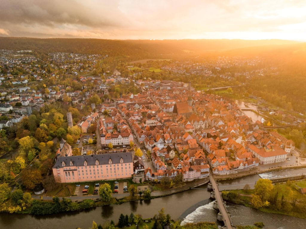 Drohnenansicht der Stadt Hann. Münden in Niedersachsen im Sonnenuntergang. Im Vordergrund ein Fluss mit einer Brücke darüber, mittig eine kleine Altstadt mit zahlreichen Fachwerkhäusern, deren rot Dächer im Licht der Sonne strahlen.