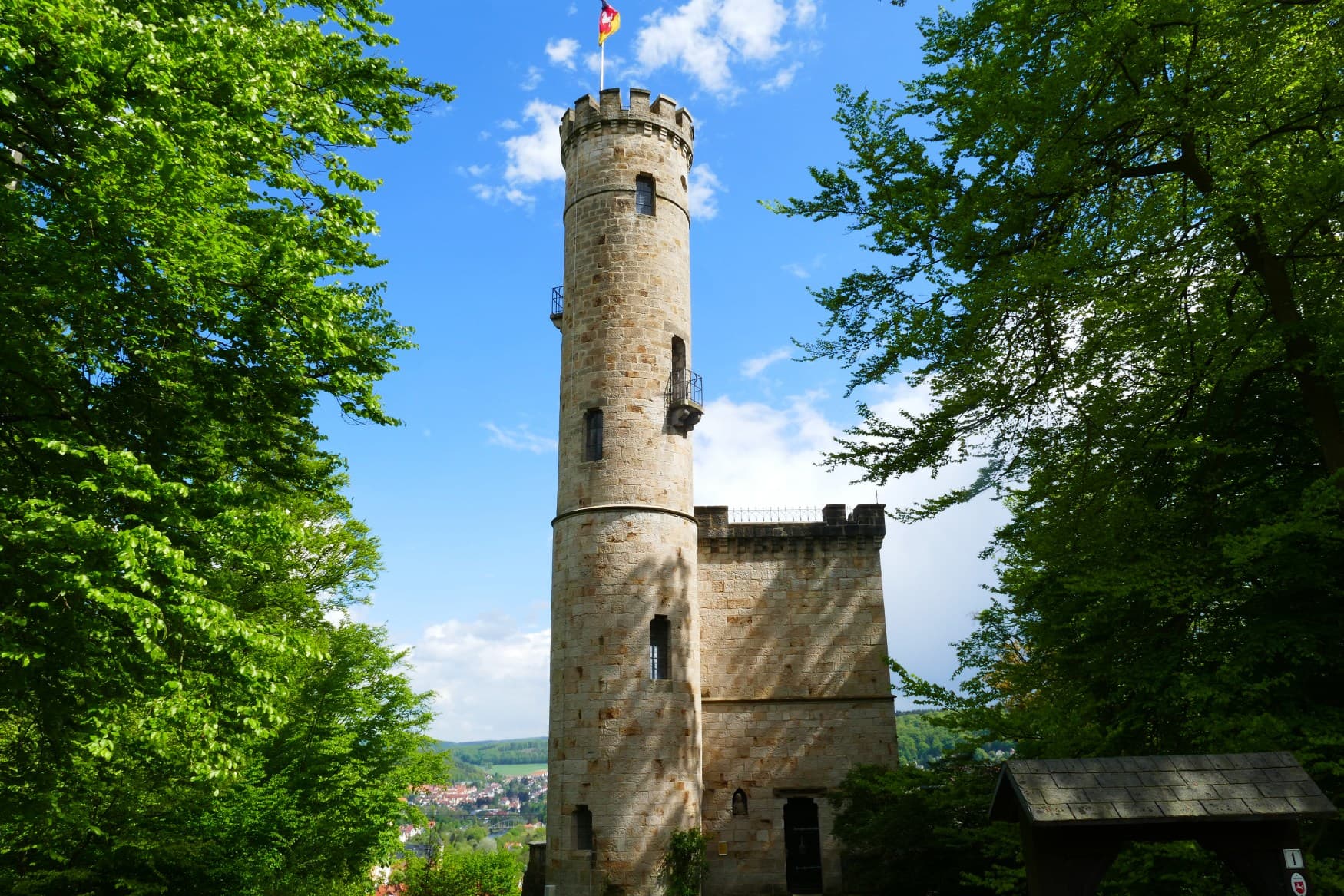 Ein neuzeitlicher Festungsbau mit einem hohen Rundturm in einem kleinen Waldstück. Dahinter liegt ein Tal mit einer Stadt.