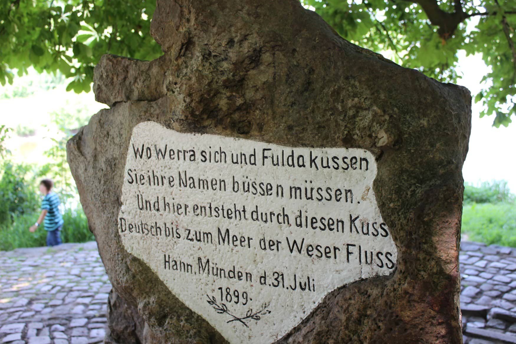 An einem Pflasterweg steht ein großer Stein. Darauf ist folgende Inschrift zu lesen: "Wo Werra sich und Fulda küssen / Sie ihre Namen büssen müssen. / Und hier entsteht durch diesen Kuss / Deutsch bis zum Meer der Weser Fluss / Hann. Münden d. 31. Juli 1899"
