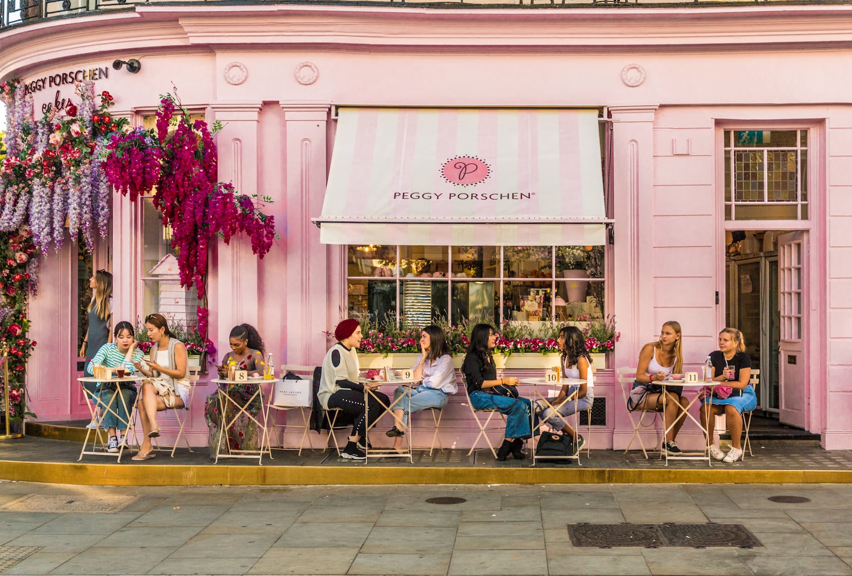Vor einem ganz in Rosa gehaltenen Café, auf dessen Markise "Peggy Porschen" prangt, sitzen mehrere Menschen je zu zweit an kleinen Tischen der Außengastronomie.