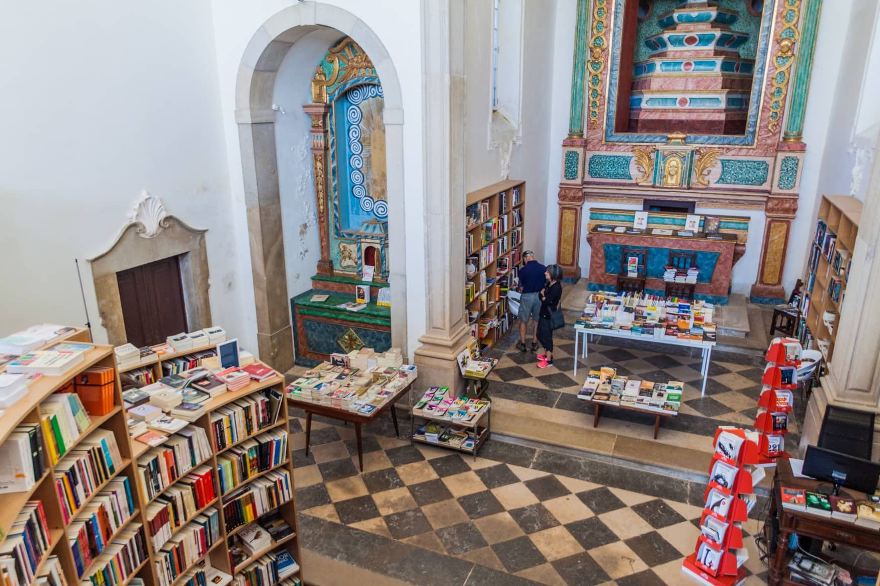 Blick von einer Empore in eine portugieseische Kirche, an deren Wänden zahlreiche Bücherregale stehen. Einige Menschen stöbern an einer Buchauslage.