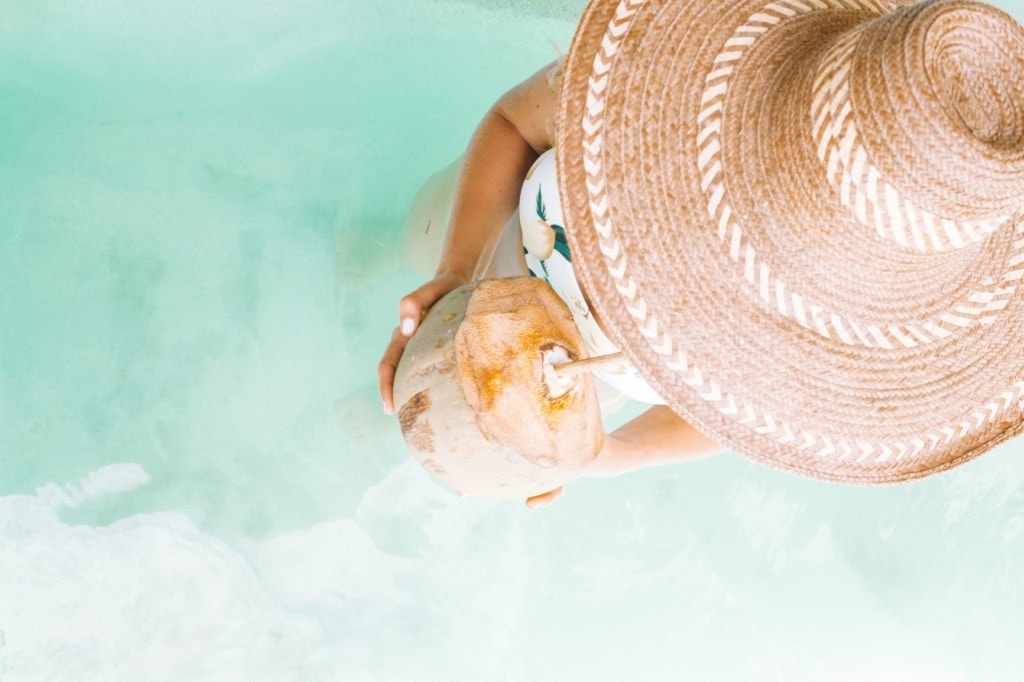 Frau mit Sonnenhut und Kokosnuss in der Hand am Strand