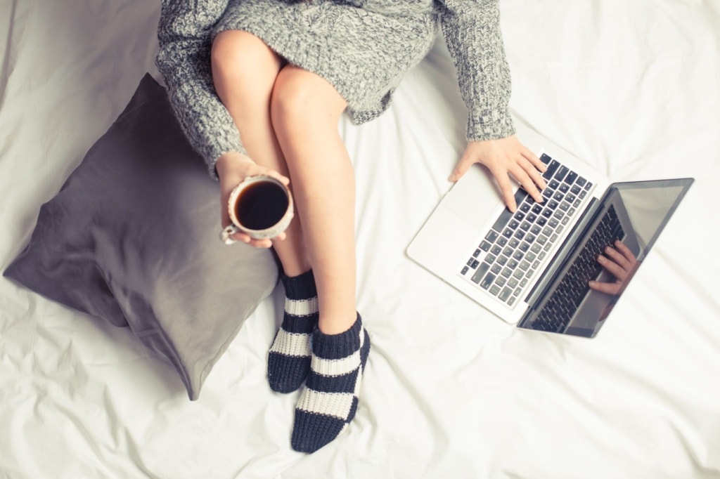 Frau sitzt im Bett, hält Tasse Kaffee in der Hand, und bedient Notebook