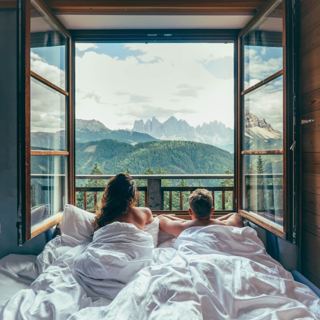 Pärchen liegt im Bett und blickt aus offenem Fenster auf die umliegende Bergkulisse