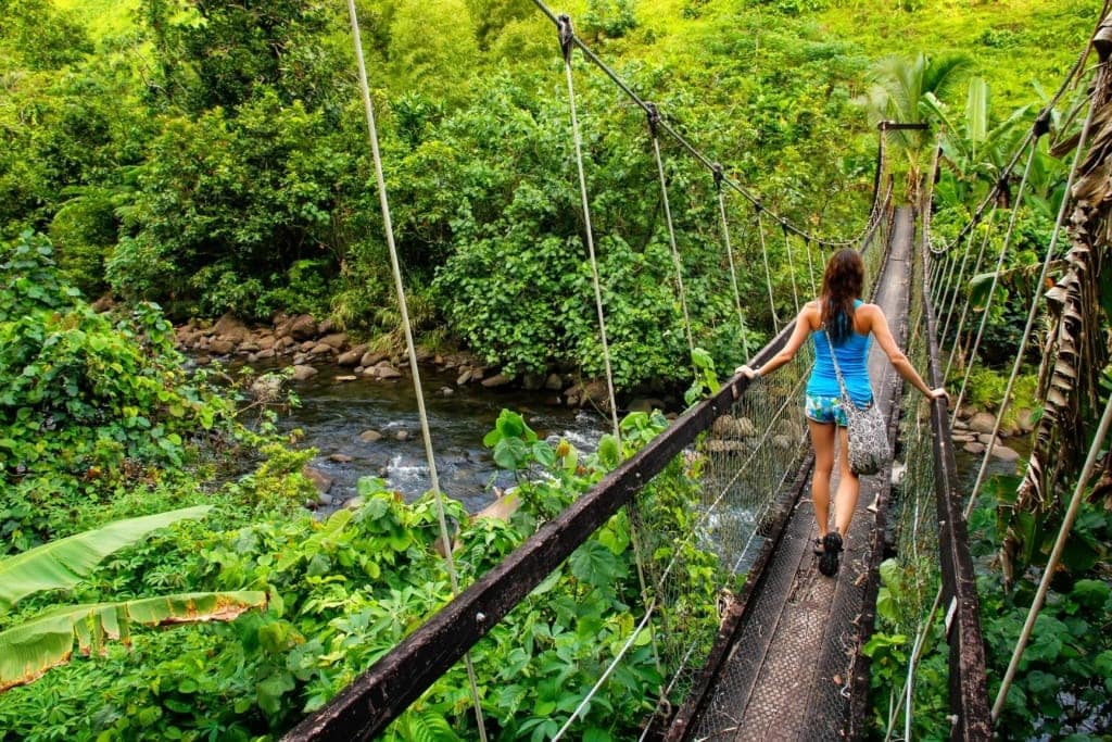 Junge Frau, die auf dem Lavena Coastal Walk über eine Hängebrücke über den Bach Wainibau auf Taveuni Island, Fidschi, geht.