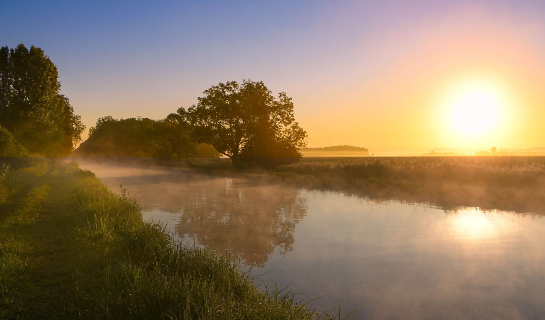 Idylle am Rhone-Kanal im Elsass; der Kanal liegt im dichten Morgendunst, am Horizont ist die Sonne bereits aufgegangen