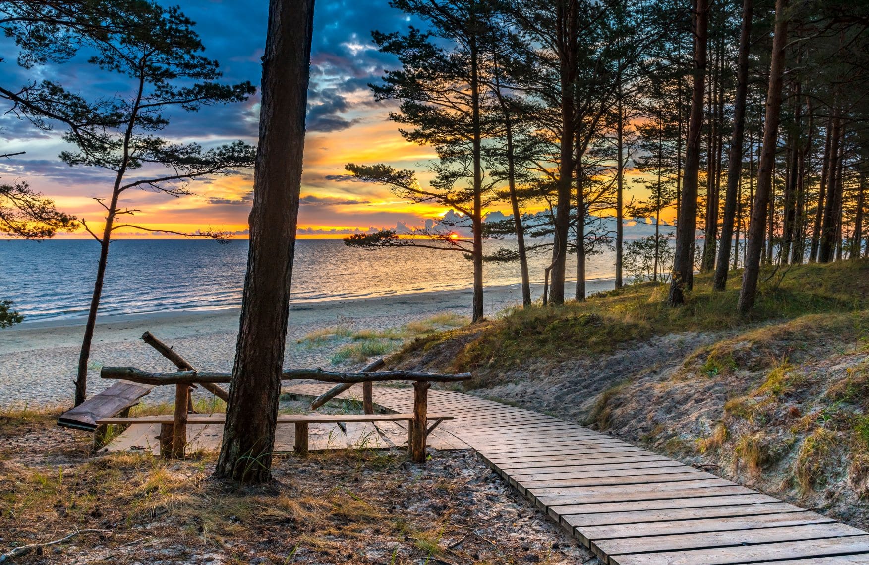 Von einer bewaldeten Anhöhe führt ein Holzsteg zu einem breiten Strand hinunter. Am Horizont geht im Meer die Sonne unter.
