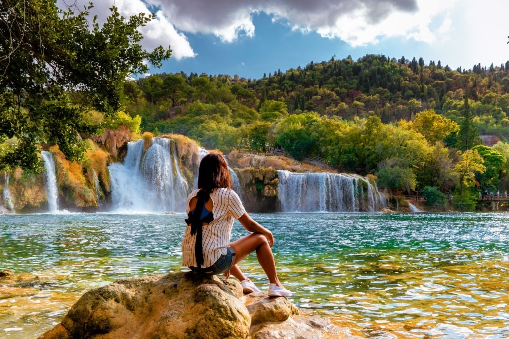 Junge Frau sitzt auf Stein und blickt auf einen Wasserfall im Nationalpark Krka, Kroatien 