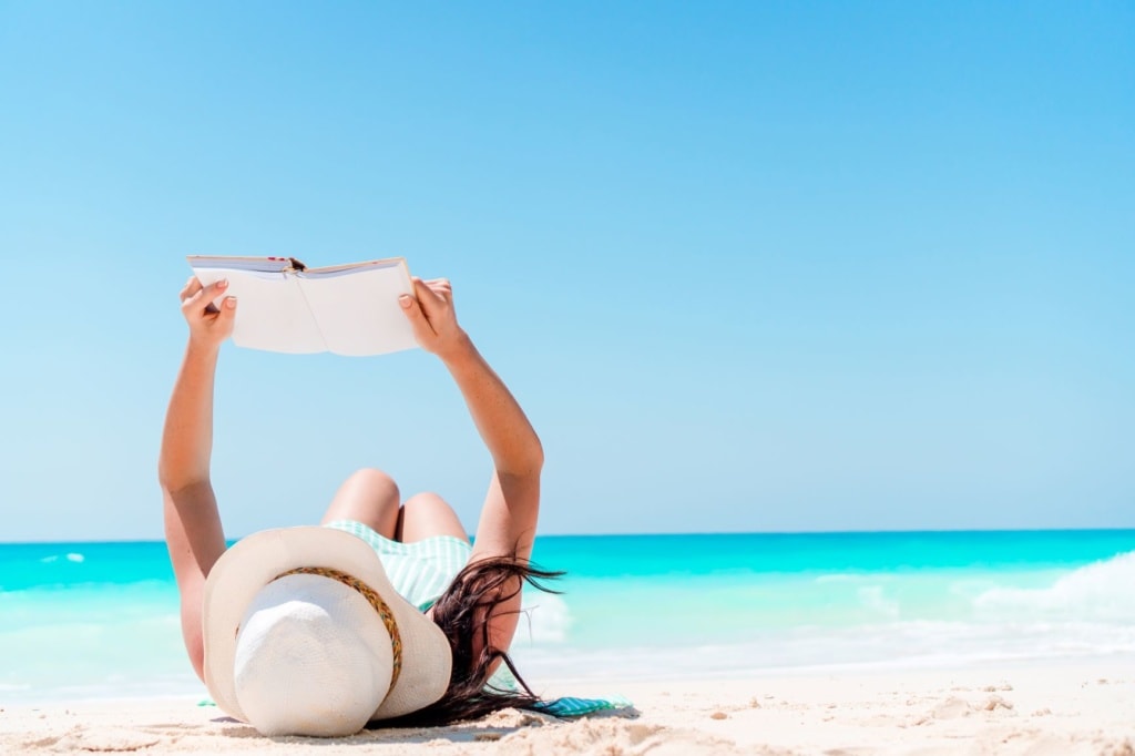 Frau liegt auf weißem Sandstrand und liest ein Buch