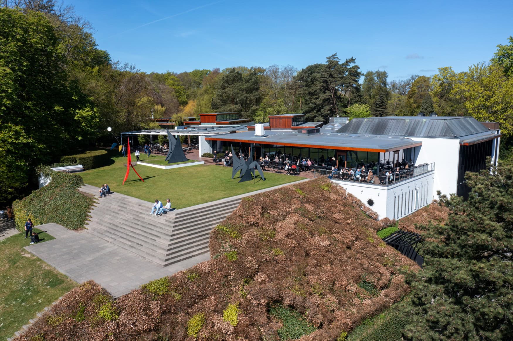 Drohnenaufnahme des Außenbereichs des Louisiana Museums of Modern Art bei Kopenhagen. Links im Vordergrund eine begrünte Terrasse, rechts dahinter ein moderner Bungalow mit breiter Glasfront, vor der Menschen in einem Café-Betrieb sitzen.