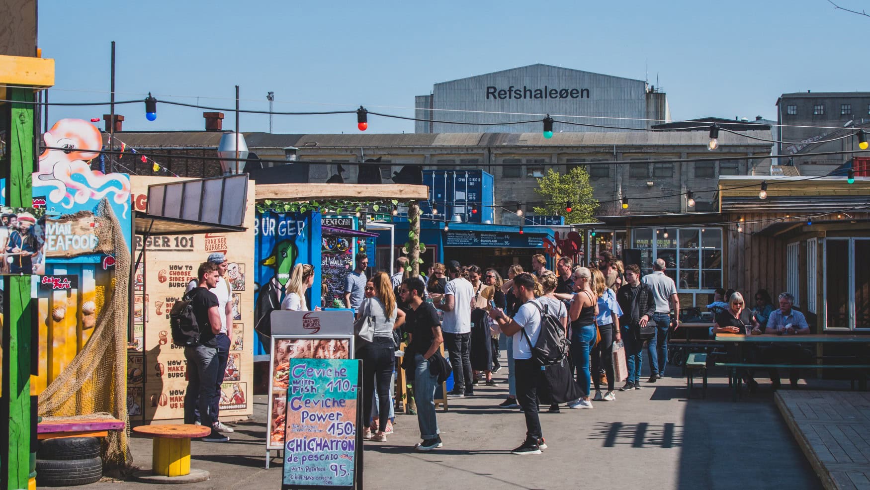 Ein Street-Food-Markt auf einer Insel in Kopenhagen. Mittig stehen einige bemalte Container, davor jeweils eine Schlange Kunden. Im Hintergrund ist ein großes Lagerhaus mit der Aufschrift "Refshaleøen" sichtbar.