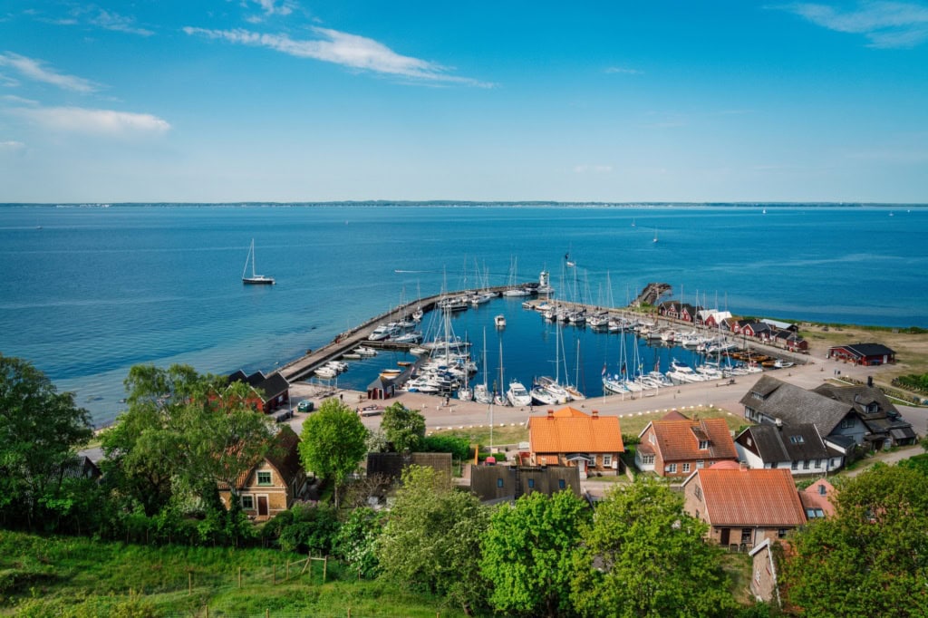 Ausblick von einem grünen Hügel auf einen kleinen Yacht-Hafen auf einer Ostseeinsel. Der Himmel über der Szene ist sommerlich-blau.