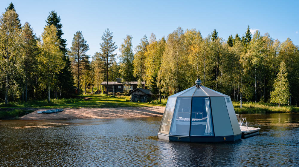 Schwimmende Glas-Lodge auf finnischem See, das Ufer dahinter dicht von Bäumen bewachsen
