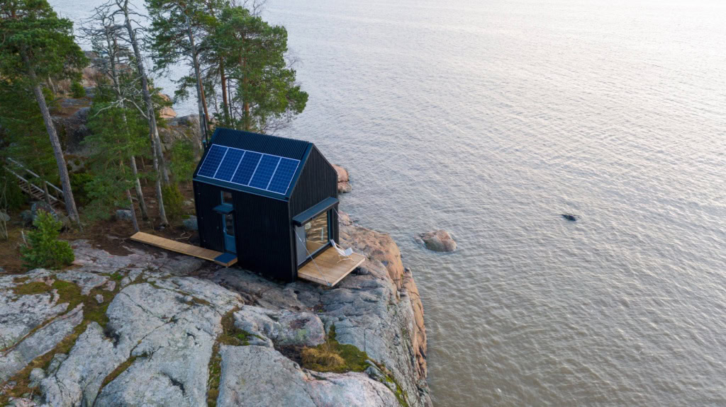 Am felsigen Ufer eines Sees steht eine kleine Lodge aus schwarzem Holz, auf dem Dach ein Solar-Panel.
