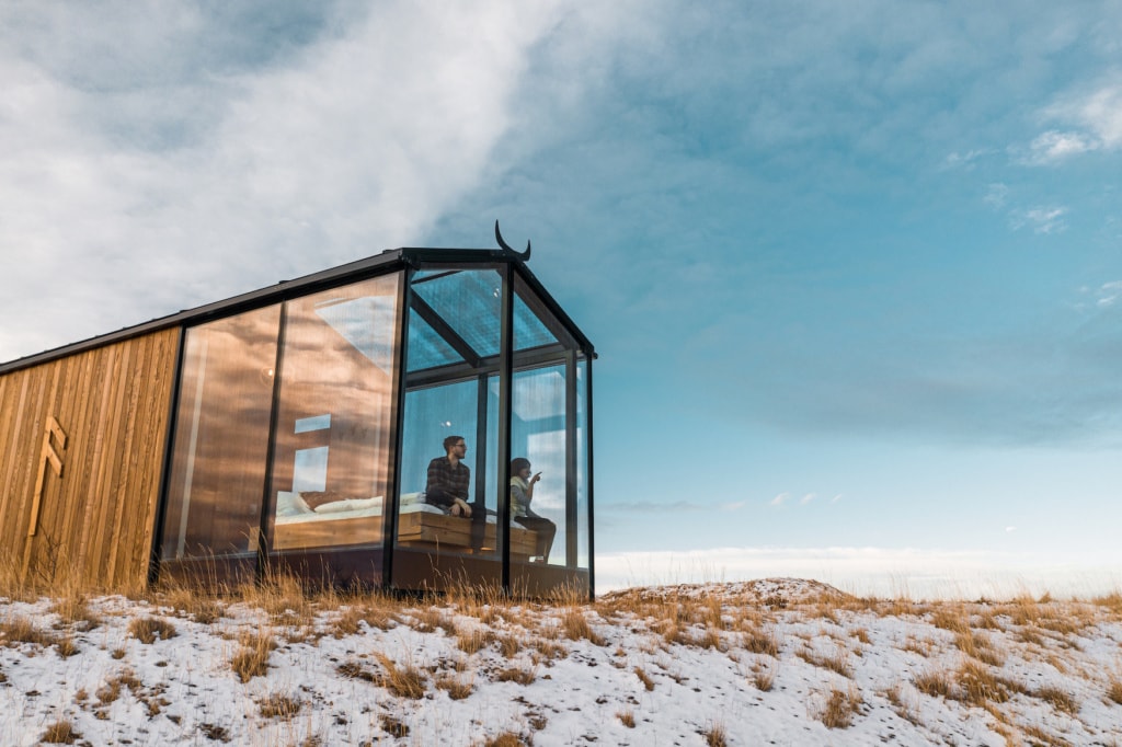 Zwei Personen sitzen im Glasanbau einer traditionellen skandinavischen Holzhütte und schauen hinaus.