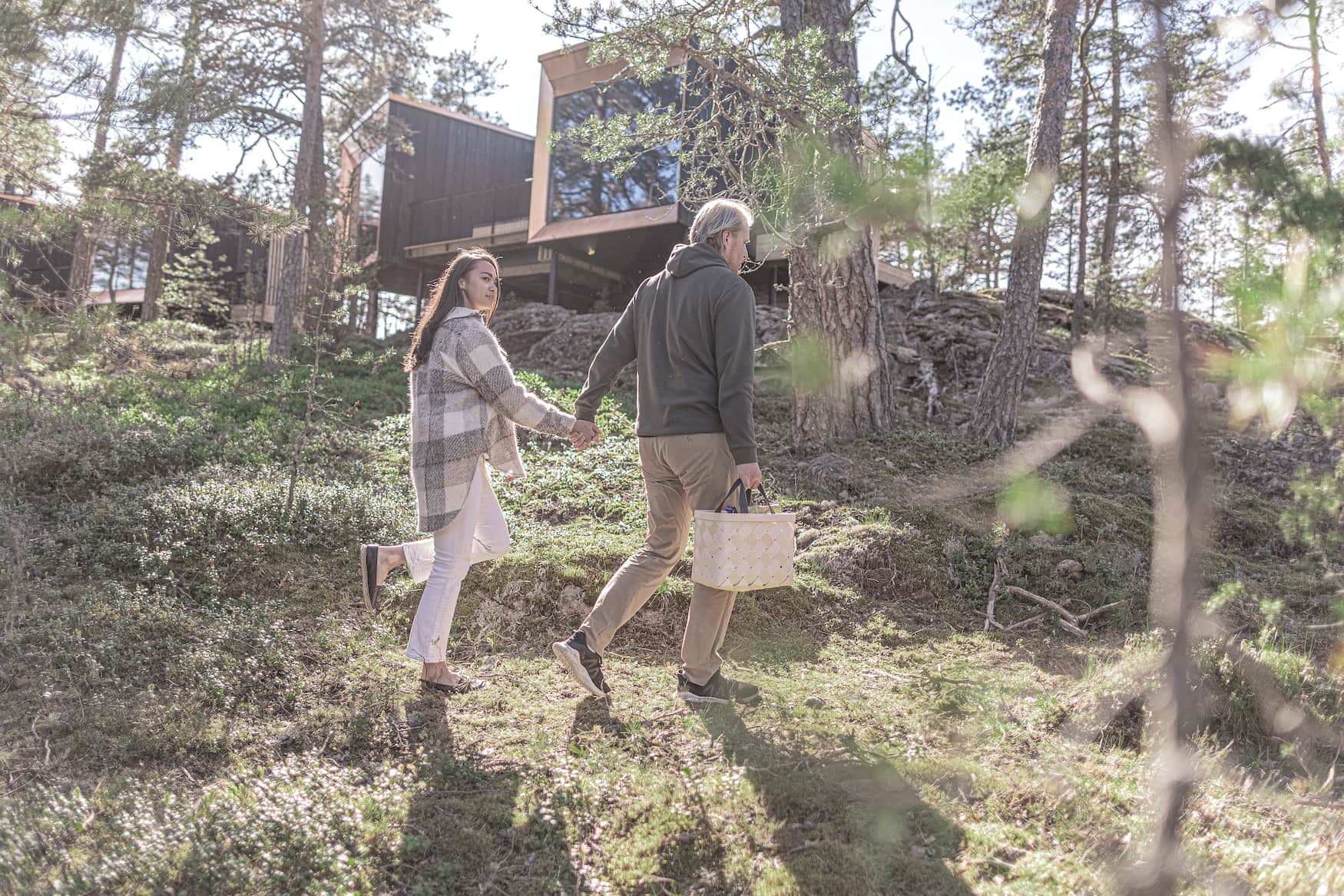 Mann und Frau spazieren durch Natur in Finnland, im Hintergrund sieht man ihr Hotel: zwei auf Stelzen stehende Holzquader mit großen Fensterfronten.