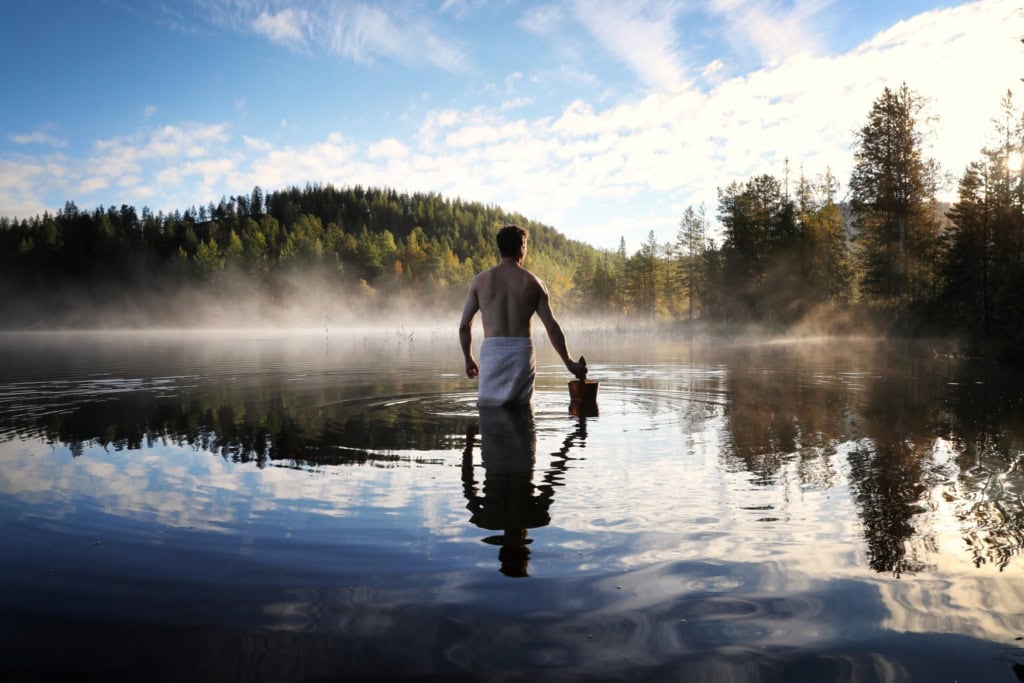 Ein Mann steht, nur mit einem Handtuch bekleidet und einem Holzeimer in der Hand, bis zu den Oberschenkeln in einem weiten See. Auf der gegenüberliegenden Uferseite beginnt ein dichter Nadelwald.