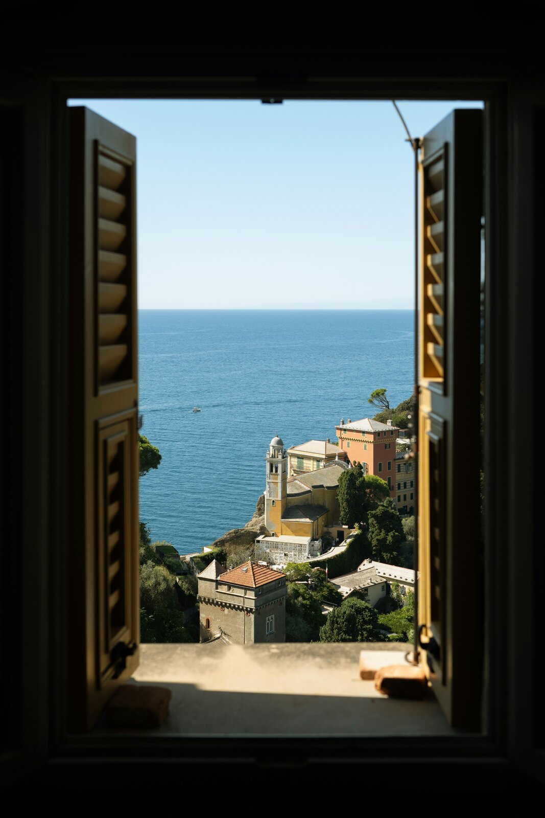 Blick durch ein geöffnetes Holzfenster mit Lamellenläden auf das Ligurische Meer und die Kirche San Giorgio von Portofino.