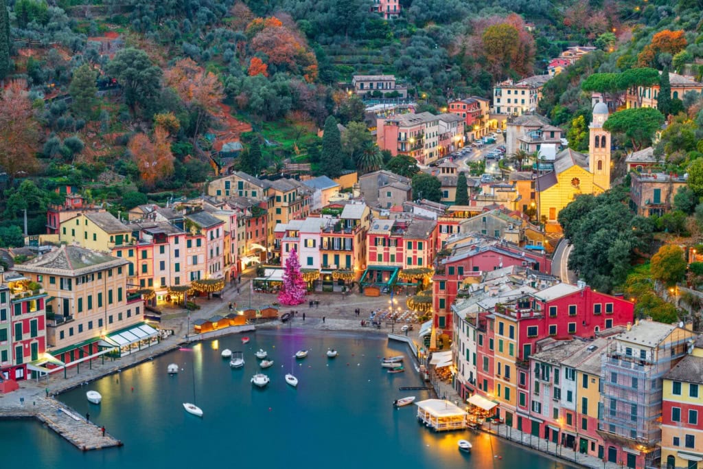 Luftaufnahme des Hafens von Portofino in der Abenddämmerung mit bunten Pastellhäusern, beleuchtetem Weihnachtsbaum auf der Piazzetta und ankernden Booten.