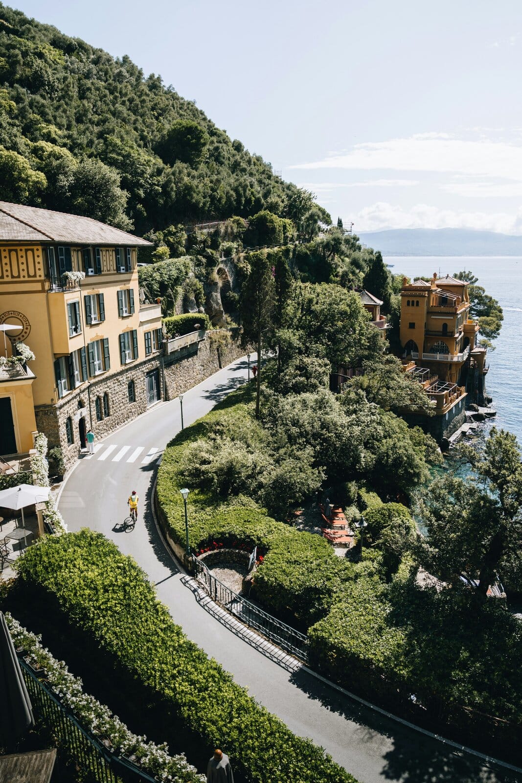 urvenreiche Küstenstraße bei Portofino mit gelben Villen, dichtem Grün und einem Radfahrer, im Hintergrund das Mittelmeer.