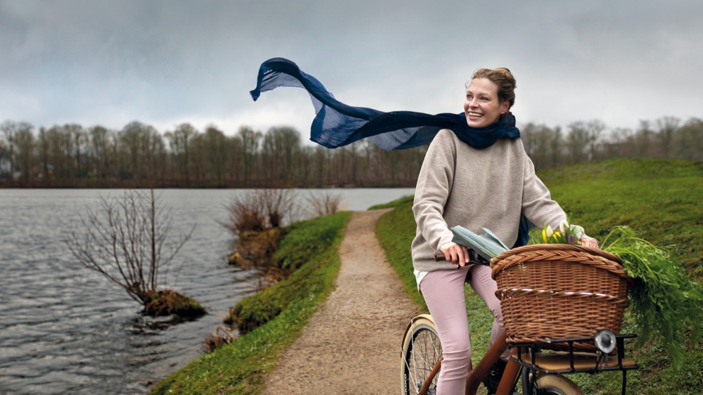 Eine Frau Fährt auf dem Fahrrad einen Weg am Ufer eines Gewässers entlang. Ihr Schal weht im Wind.