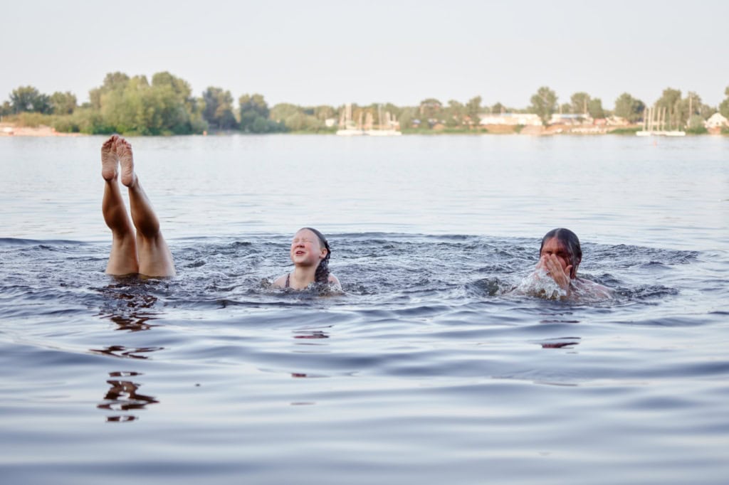 Drei Personen schwimmen in einem See, von einer sind nur die Füße und Beine zu sehen, die gerade aus dem Wasser herausgestreckt sind.