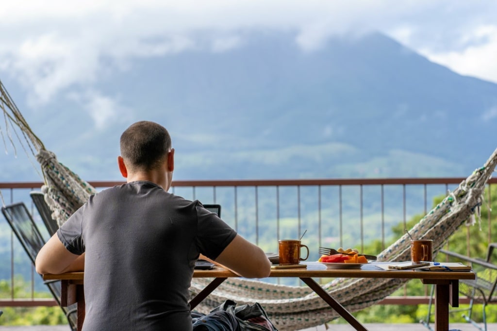 Mann sitzt auf Balkon in Costa Rica, frühstückt und arbeitet am Notebook