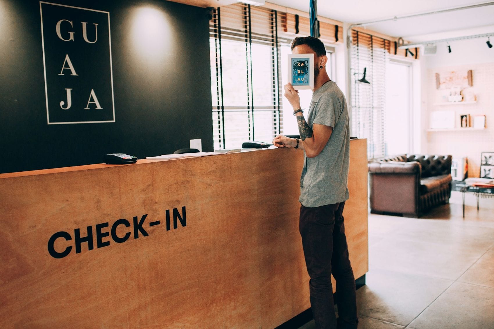 Dünner Mann, Typ Hipster, steht am Check-in-Tresen eines Hotels und hält sich ein Zettel vors Gesicht 