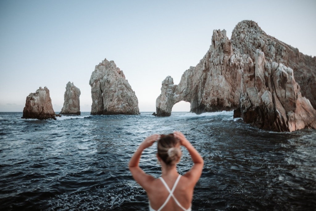Blode Person fasst an ihre Haare und blickt auf das Meer. In der Ferne sieht man den el arco de cabo san lucas aus dem Meer ragen.