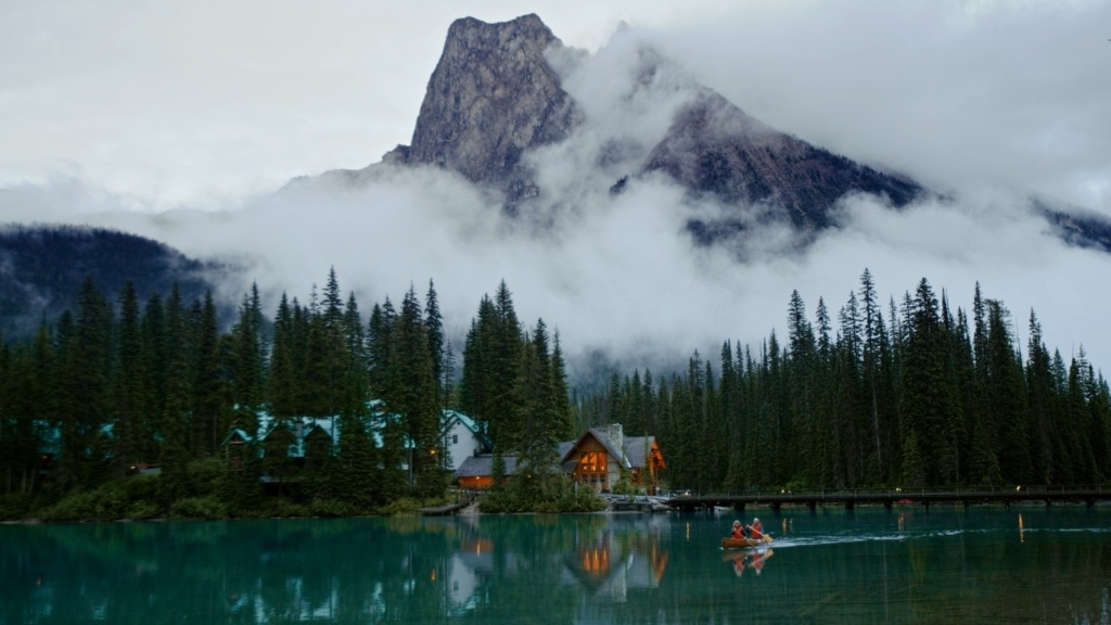 Paar fährt Kanufahren auf dem Smaragdsee im Yoho National Park in British Columbia, Kanada am frühen Abend, im Hintergrund leuchtende Berge und Lichter der Emerald Lake Lodge.