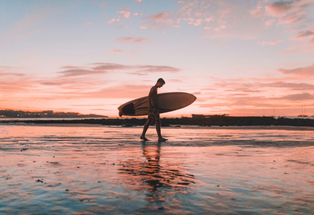 Mann mit Surfbrett bei Sonnenuntergang am Strand